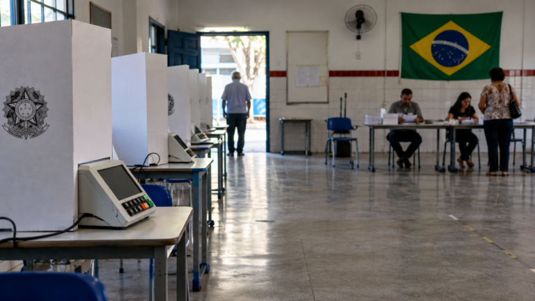 Urnas eletrônicas em seção de votação vazia, com eleitores ao fundo
